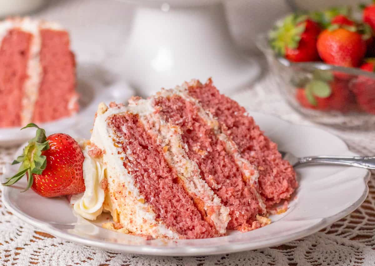 Strawberry Crunch Cake | thewendthousekitchen.com A slice of strawberry crunch cake with a fresh strawberry on a white plate with strawberries in the background.