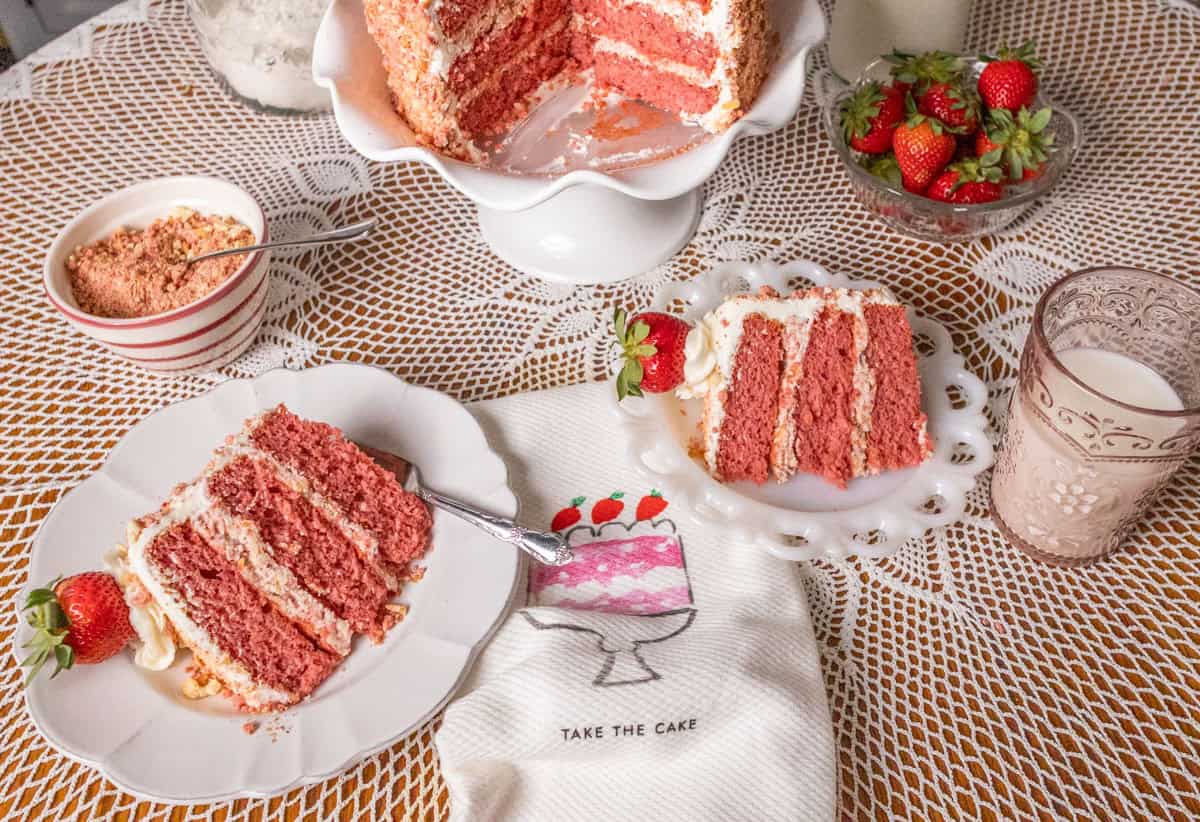 Strawberry Crunch Cake | thewendthousekitchen.com Two slices of strawberry crunch cake on white plates with the cake on a white pedestal in the background. A glass of milk and a bowl of strawberries on the table. A dish towel with a pink cake that says "take the cake".